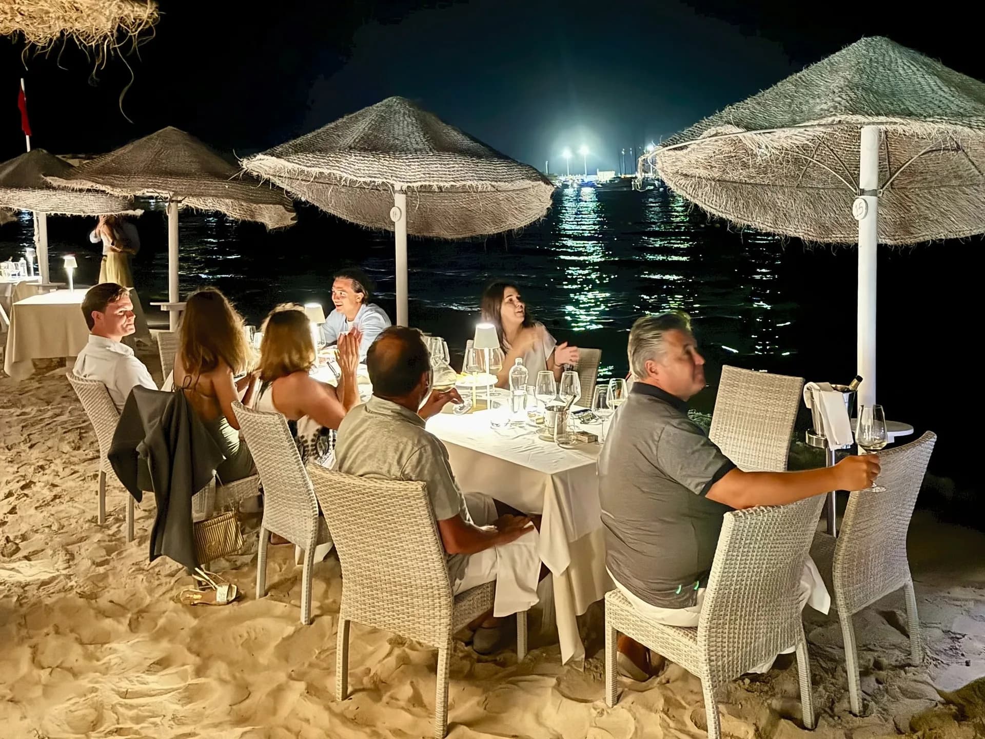 Gruppo di ospiti a un tavolo con tovaglia bianca sulla sabbia di sera, sotto ombrelloni di paglia intrecciata, con calici, lampade da tavolo e il mare e le luci del porto illuminati sullo sfondo