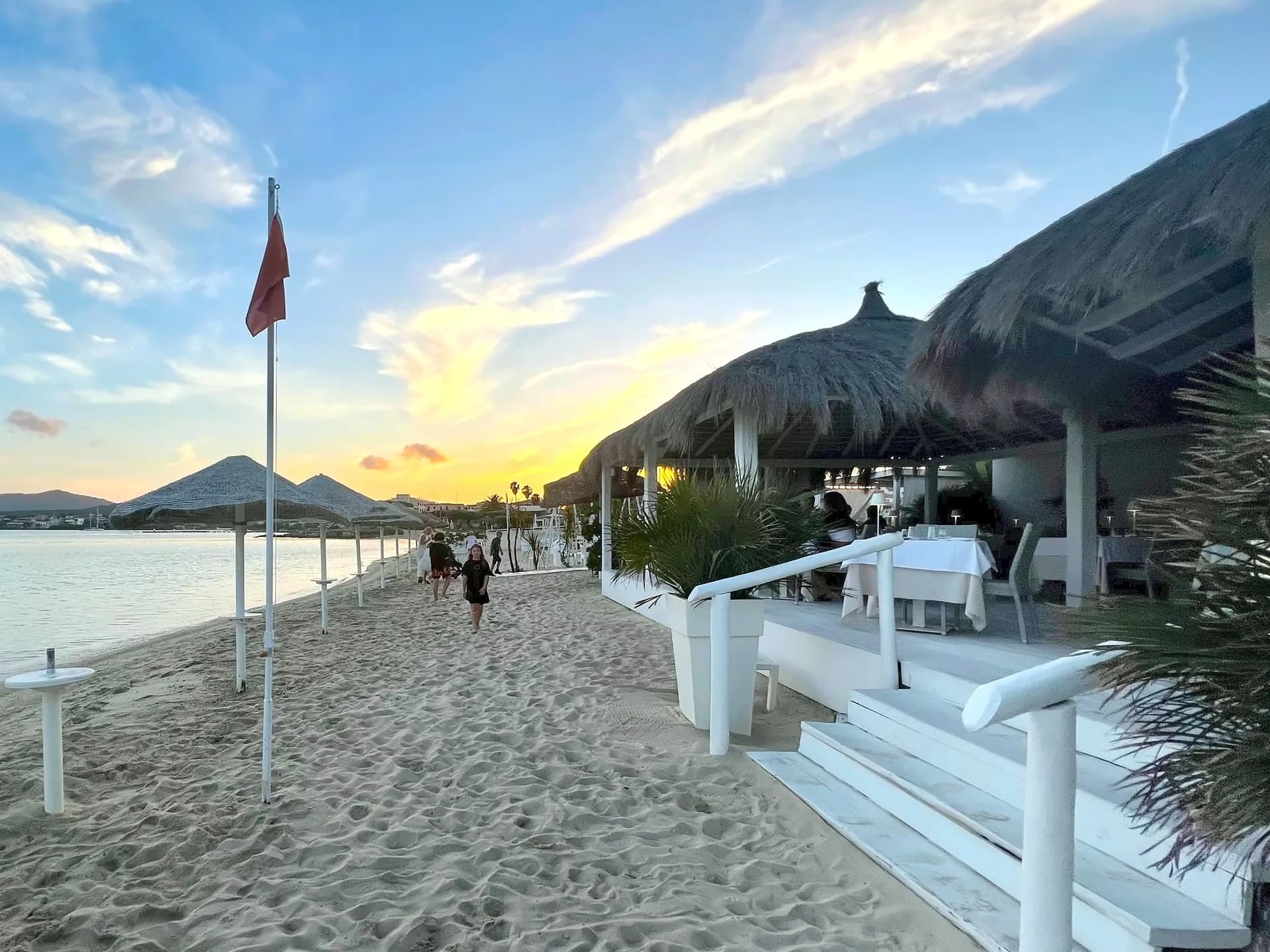 Thatched beachfront restaurant with white-clothed tables beside a sandy shore at sunset, with straw parasols, a red flag and people strolling along the calm bay