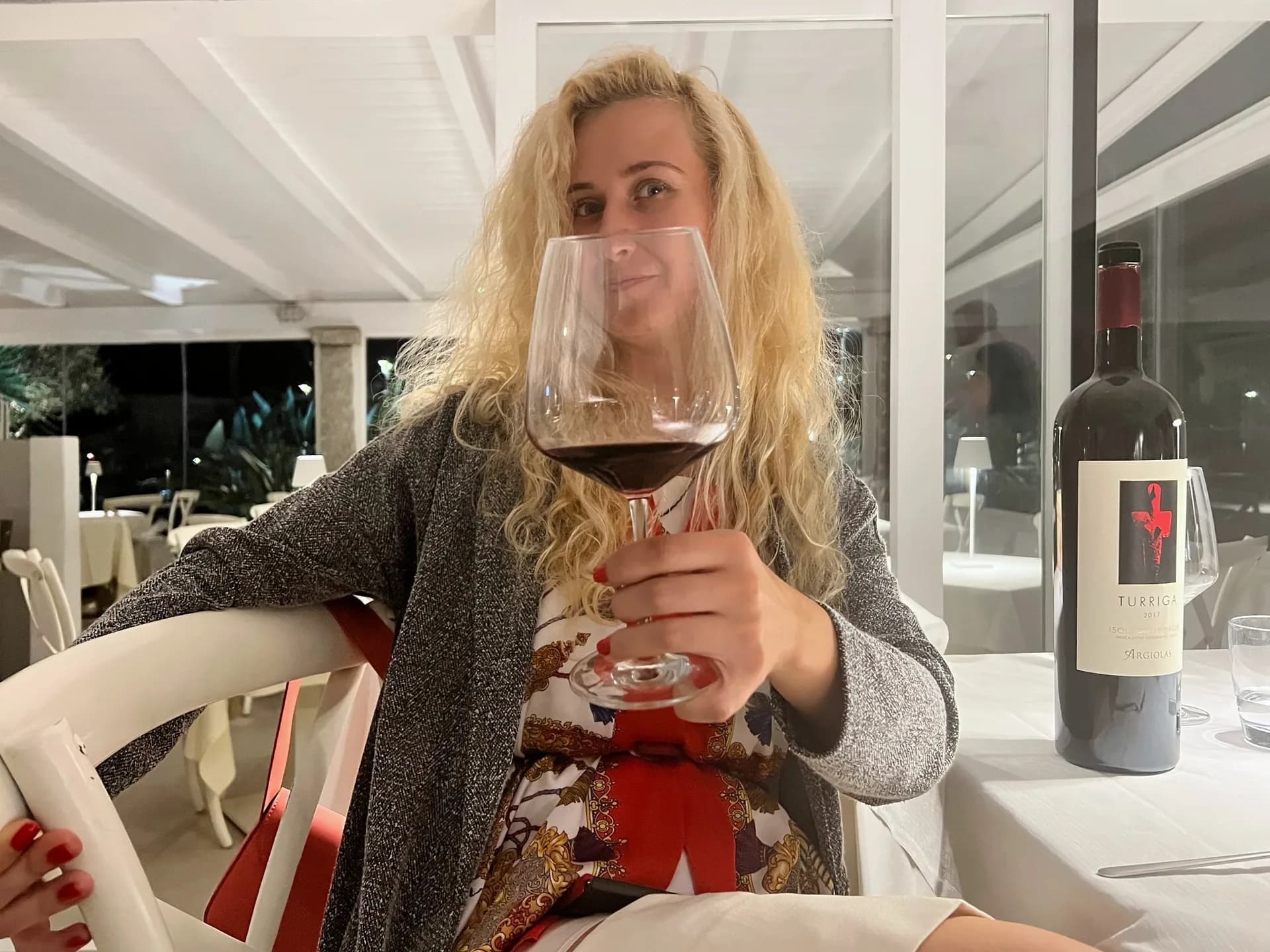 Donna con capelli biondi ricci che tiene davanti al viso un grande calice di vino rosso a un tavolo con tovaglia bianca, con accanto una bottiglia con etichetta Turriga in un ristorante luminoso di sera.