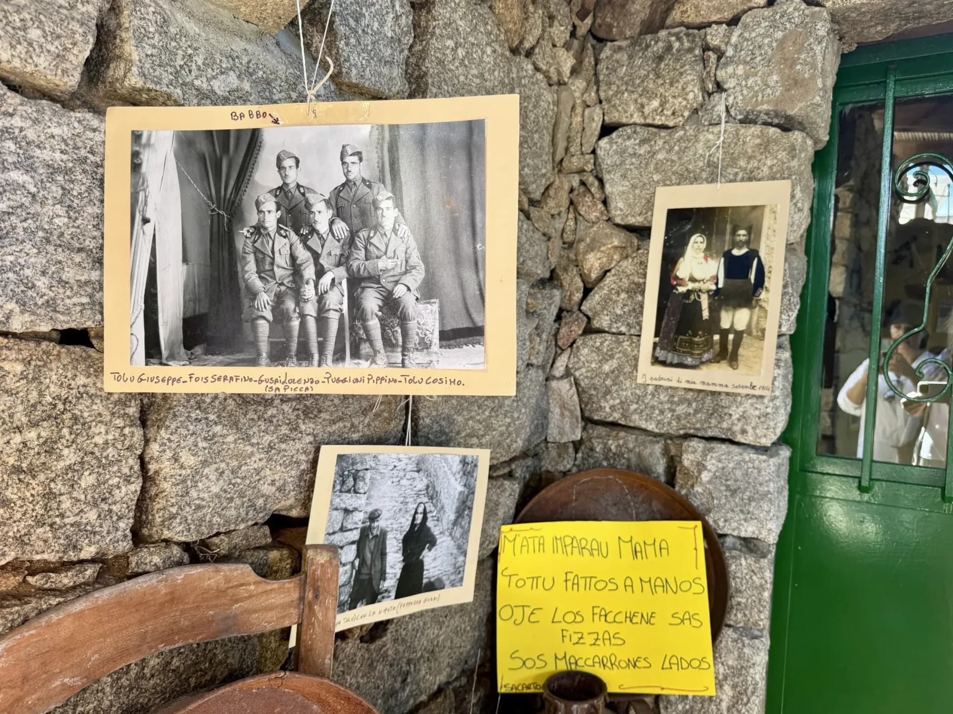 Old family photographs and a handwritten sign in Sardinian on a stone wall beside a green door in Lollove