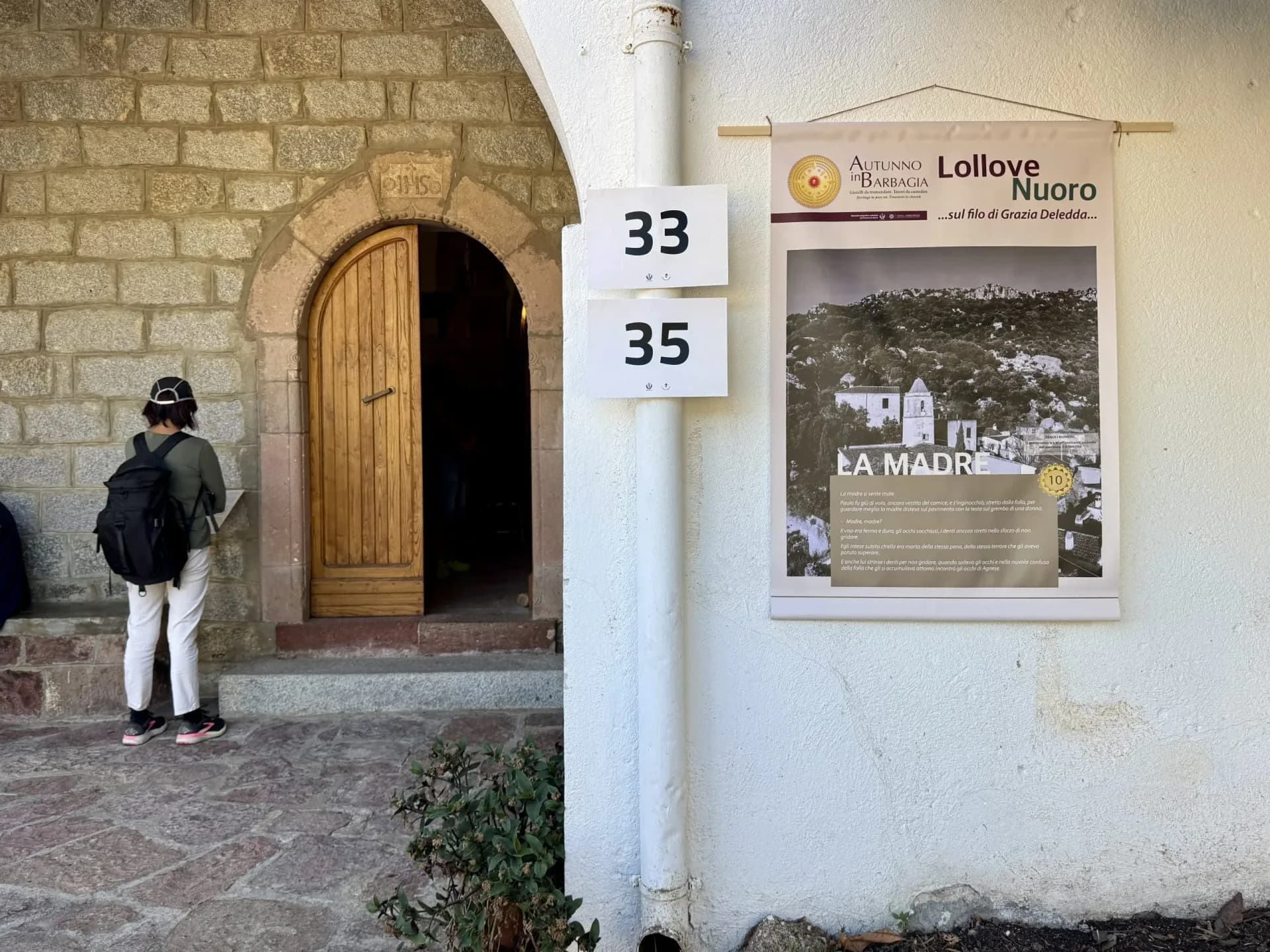 Visitor at an open wooden door of a stone house in Lollove, beside an Autunno in Barbagia banner about Grazia Deledda's novel La madre