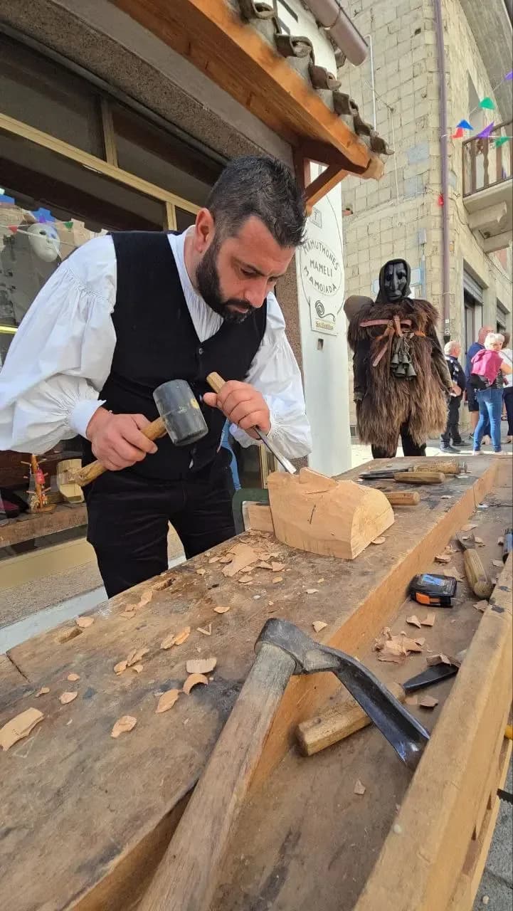 Craftsman carving a wooden mask with mallet and chisel at a workbench in Mamoiada, a Mamuthone in dark sheepskin standing behind