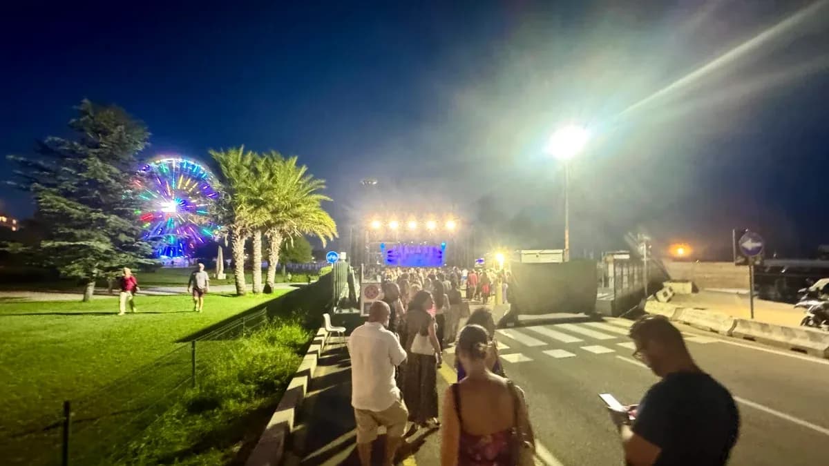 Foule marchant le long du front de mer du Molo Brin à Olbia, de nuit, vers une scène de concert éclairée, avec la grande roue illuminée derrière les palmiers