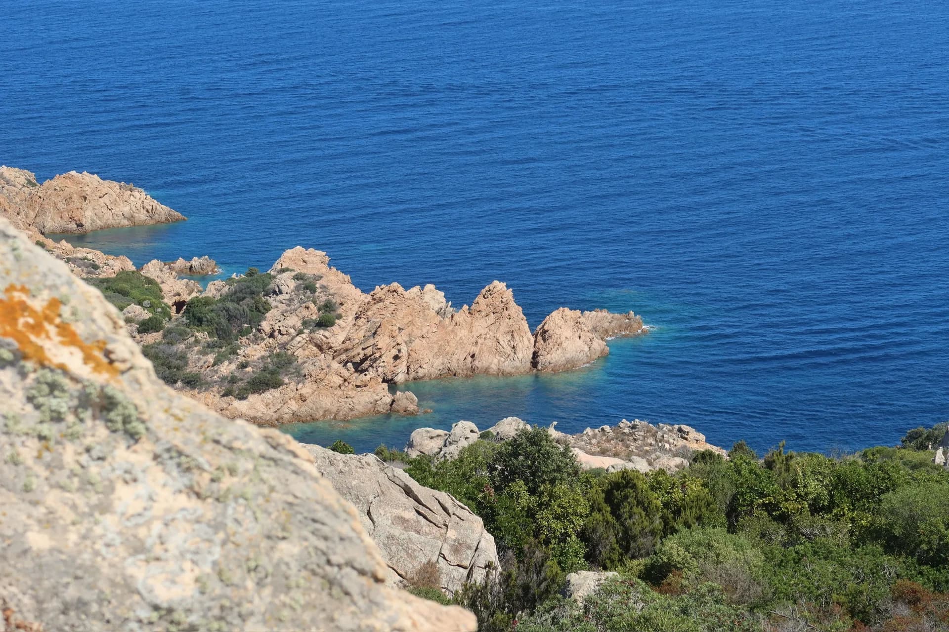 View from a rocky clifftop over jagged granite outcrops and green scrub along the Sardinian coastline, with clear turquoise shallows meeting deep blue sea.