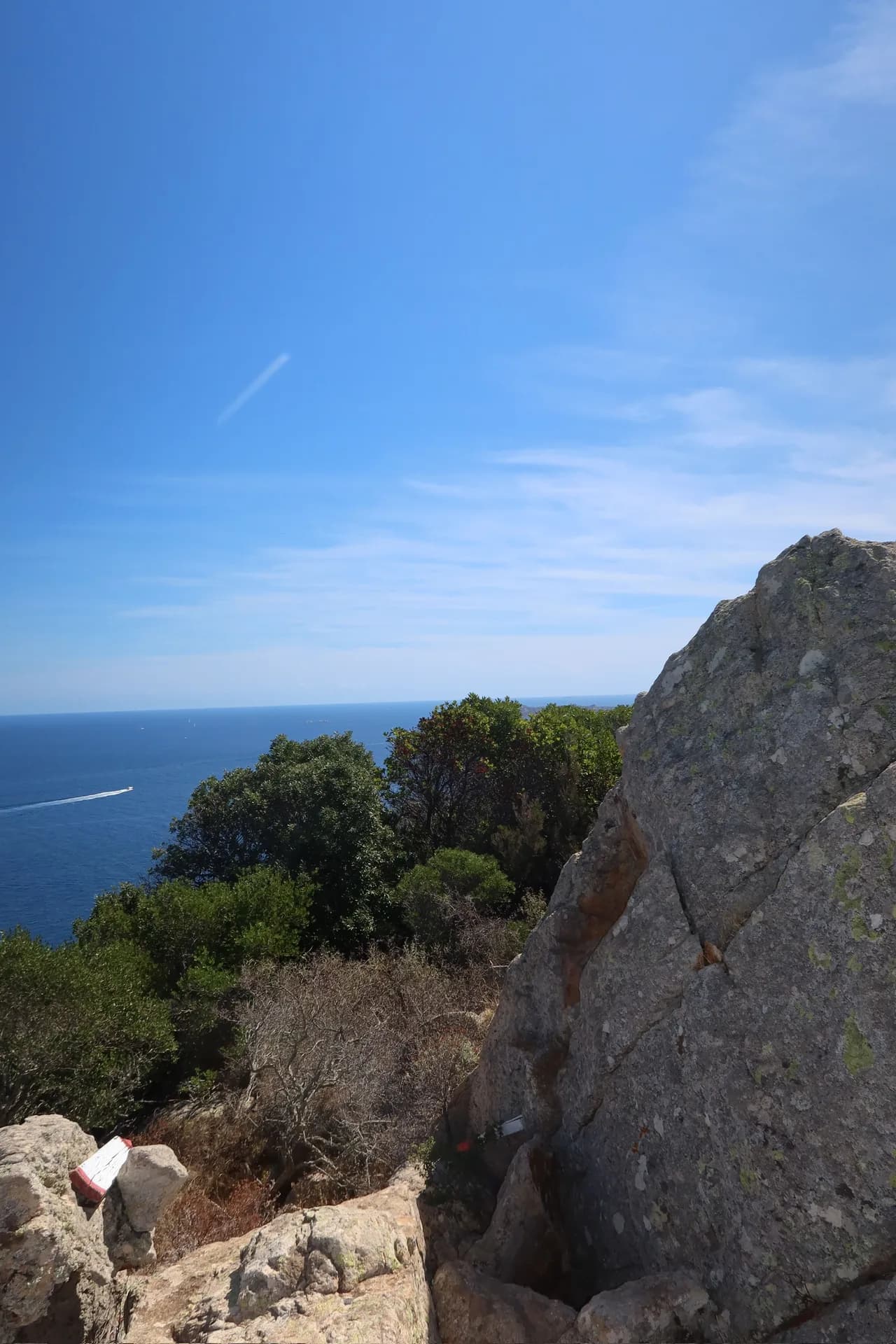 Granite boulders and Mediterranean scrub on the Pevero Health Trail, with a boat crossing the blue sea below Monti Zoppu.