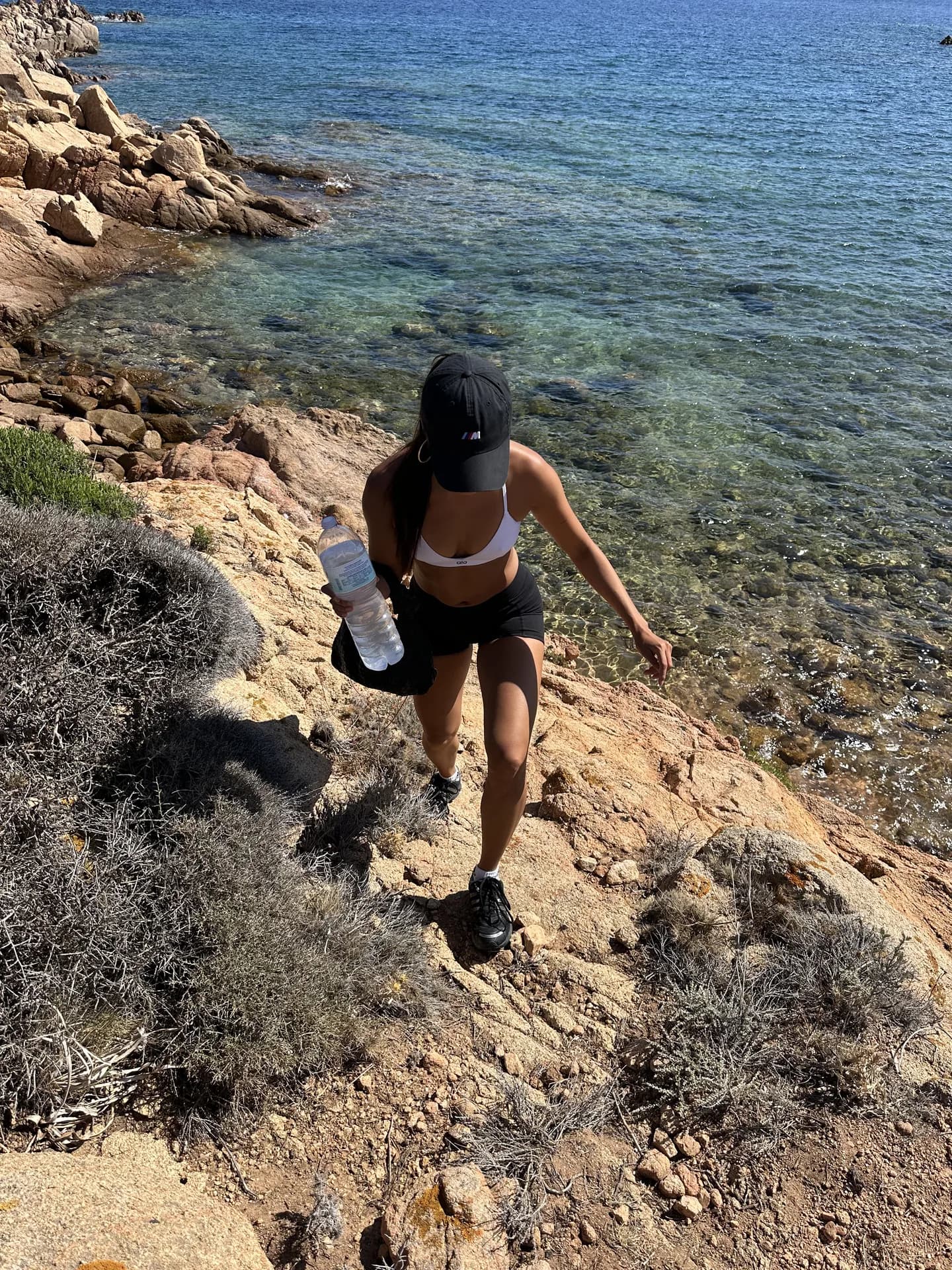 Hiker in sportswear carrying a water bottle climbs a rocky coastal trail beside the clear turquoise sea on the Pevero Health Trail, Sardinia.