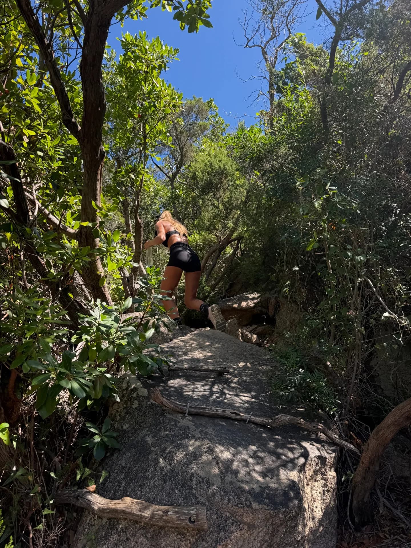 Hiker climbing a smooth granite boulder on a shaded woodland path during the Pevero Health Trail hike up Monti Zoppu, Sardinia.