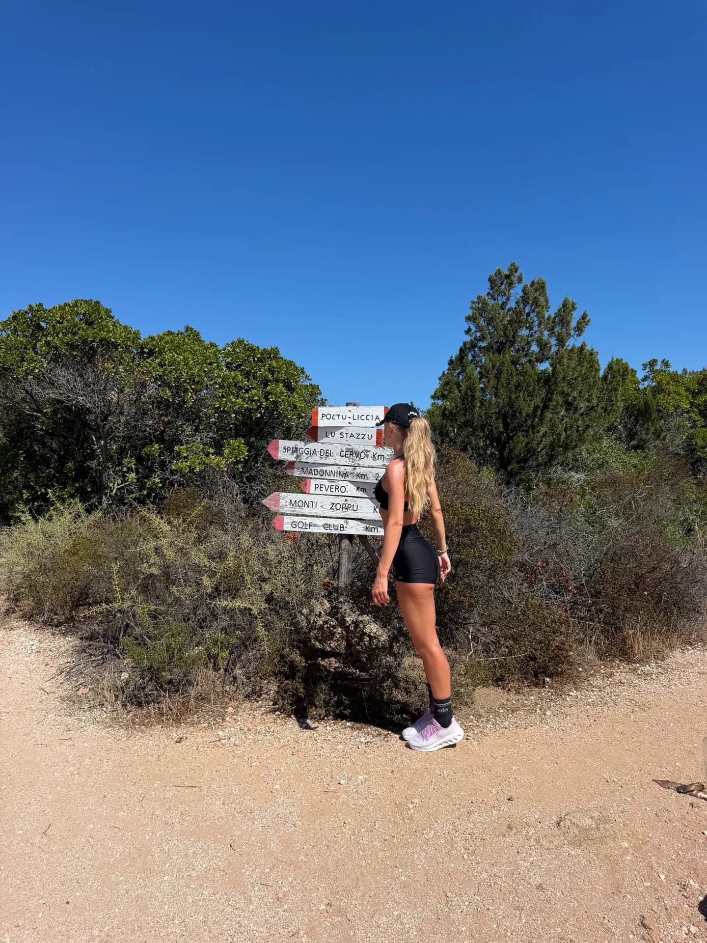 Hiker pausing beside a wooden trail signpost pointing to Monti Zoppu, Pevero, Golf Club and other destinations, along a sandy path through Sardinian scrubland under clear blue sky.
