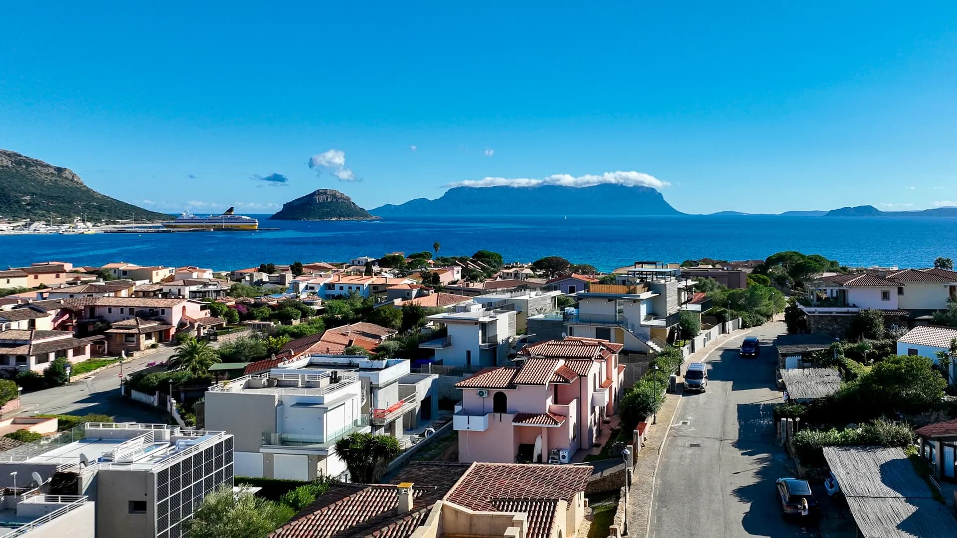 Aerial view of Golfo Aranci's modern villas and rooftops near AZULIS Villas Dumas, with Tavolara island and a ferry on the blue sea beyond.