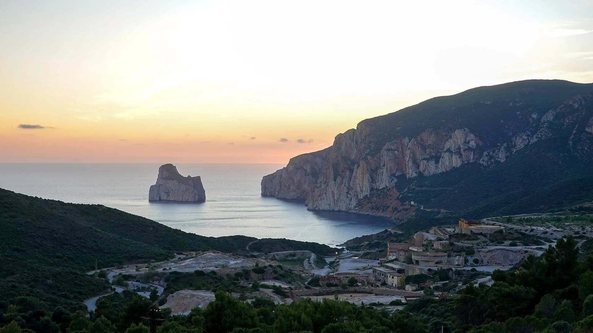 Tramonto su una baia calma con un grande faraglione al largo, ripide scogliere calcaree a destra e vecchi edifici minerari industriali tra le colline verdi in primo piano