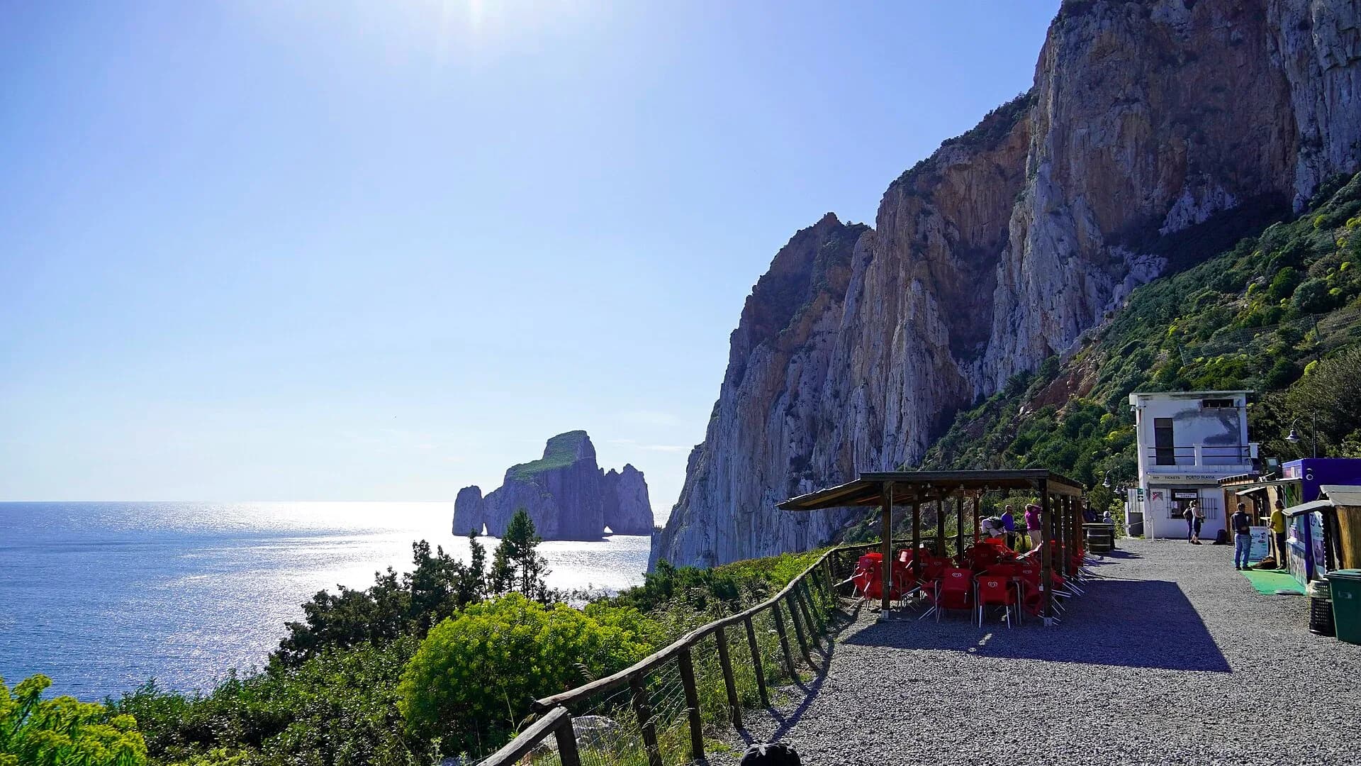 Sentiero di ghiaia ai piedi di un'alta scogliera a picco, con biglietteria, chioschi e un caffè all'ombra con sedie rosse, affacciato su un mare soleggiato e su un faraglione roccioso