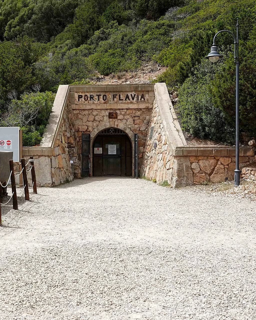 Ingresso di una galleria in pietra con una porta scura ad arco e la scritta "Porto Flavia" sopra, incastonato in un pendio verde accanto a uno spiazzo di ghiaia, un lampione e una barriera di corda.