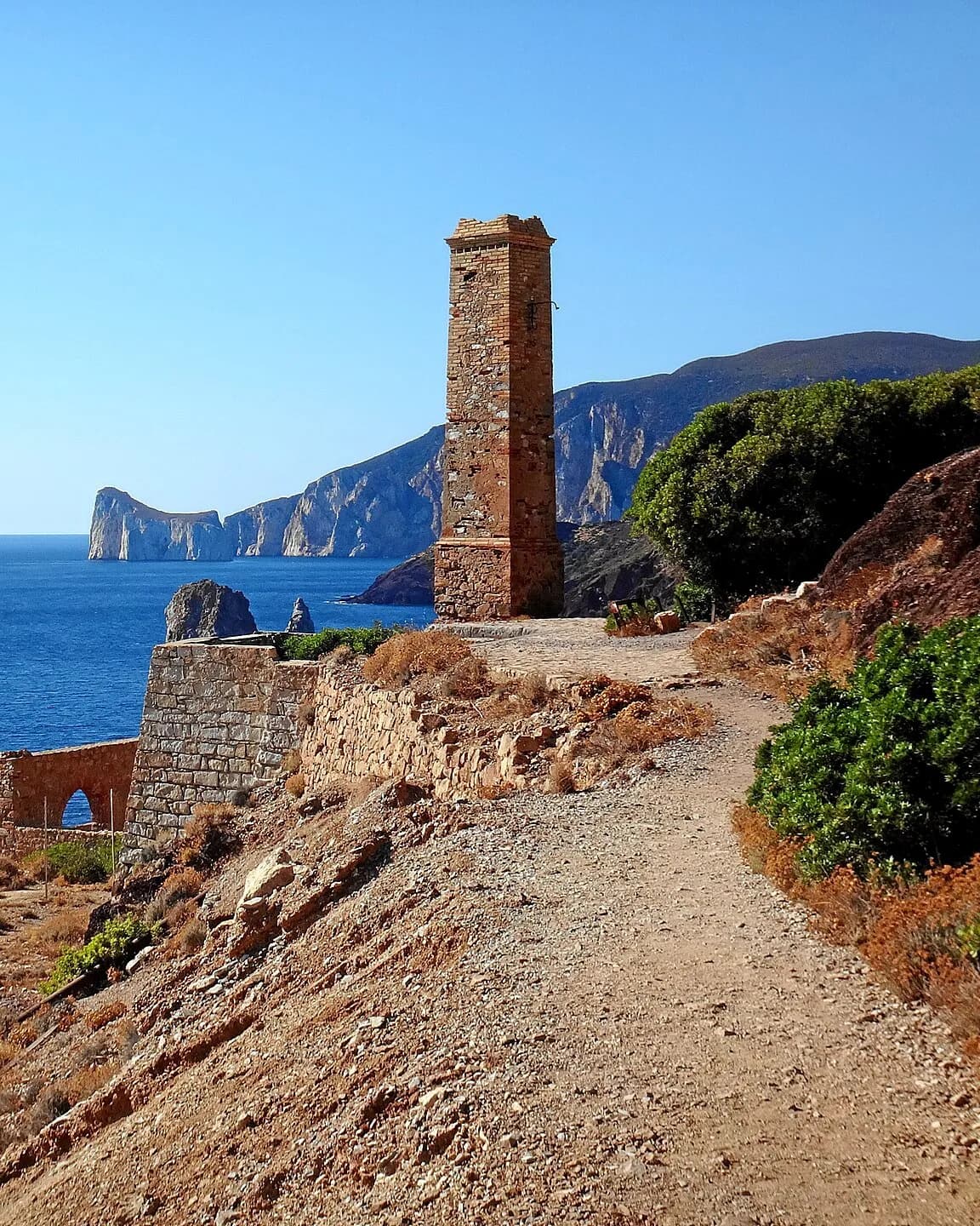 Sentiero sterrato lungo un pendio costiero che porta a un'alta torre in mattoni e pietra accanto a vecchi muri in pietra, con un grande faraglione bianco e ripide scogliere sopra il mare blu intenso
