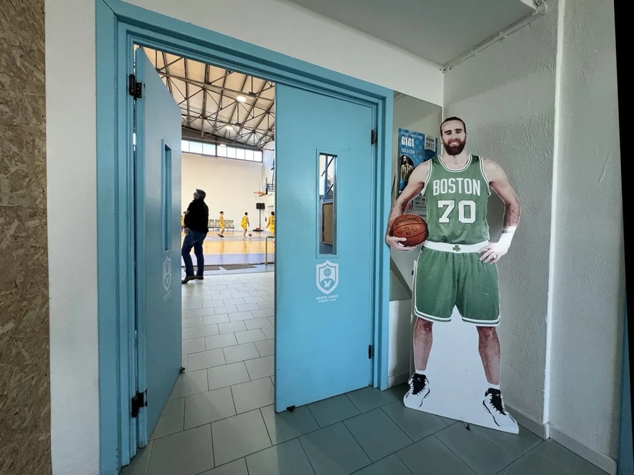 Levensgrote kartonnen figuur van een bebaarde basketballer in een groen Boston-tenue met nummer 70 naast openstaande lichtblauwe deuren, met jonge spelers op het veld van een sporthal erachter.