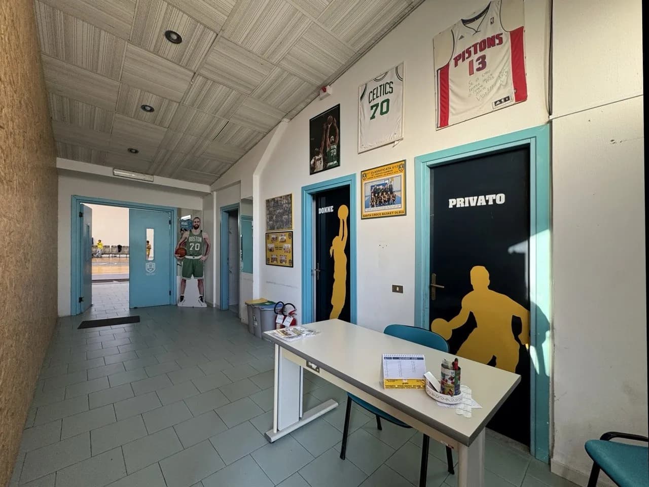 Gang van een sporthal met een bureau, basketbalsilhouetten op donkere deuren met de opschriften "Donne" en "Privato", ingelijste shirts van de Celtics en Pistons, teamfoto's en een kartonnen Boston-figuur.