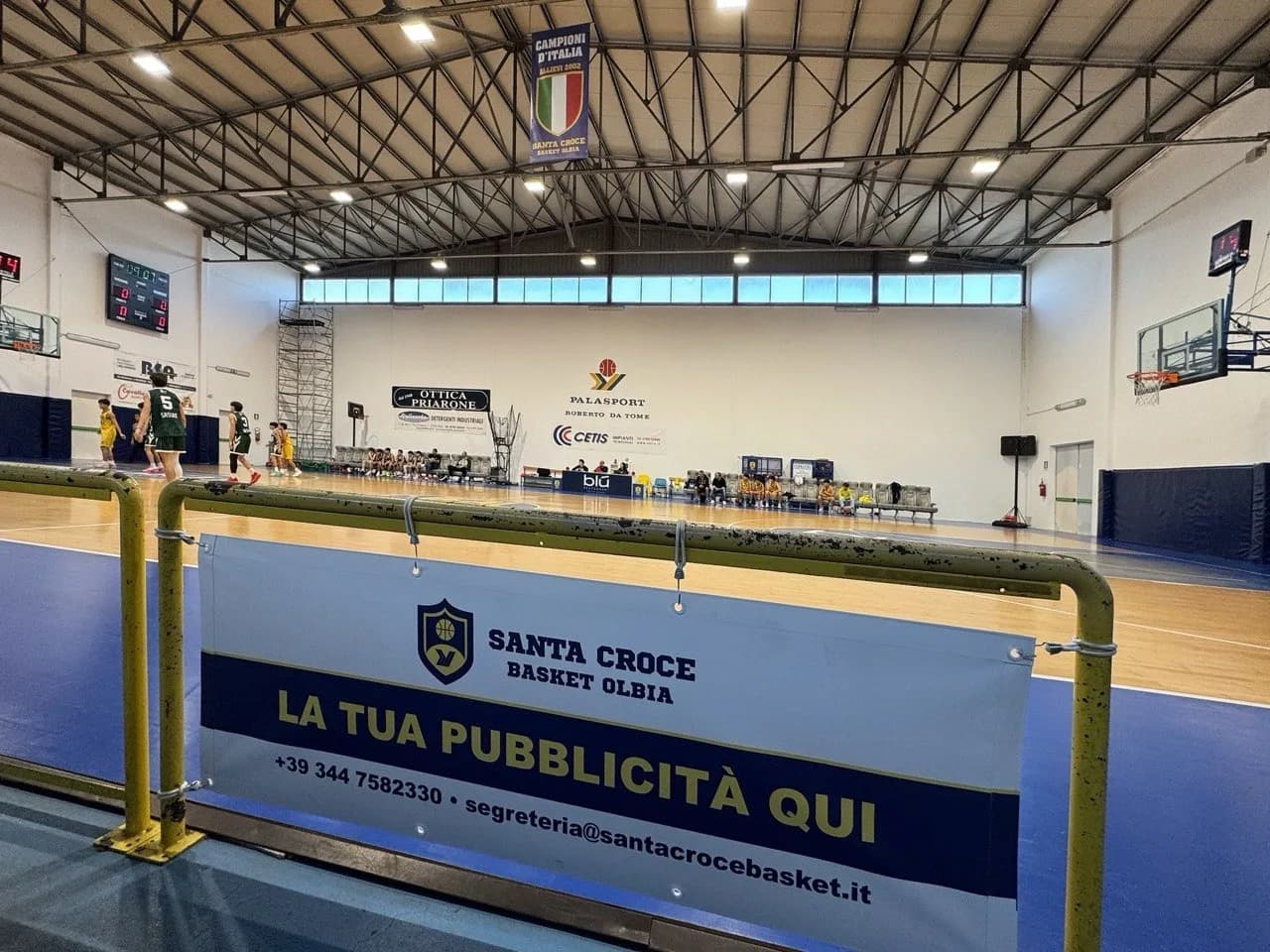 Overdekt basketbalveld tijdens een jeugdwedstrijd, met een reclamebanner van Santa Croce Basket Olbia op een gele reling op de voorgrond en een kampioenswimpel aan het dak.