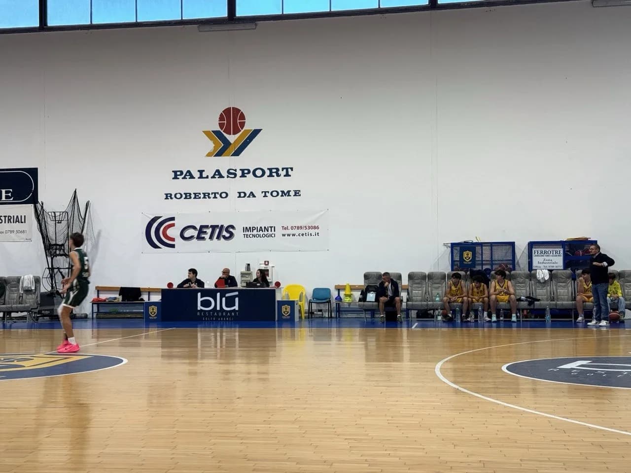 Overdekt basketbalveld in Palasport Roberto Da Tome, met een speler in het groen op de houten vloer, officials aan de scorerstafel en jongeren in geel tenue op de bank