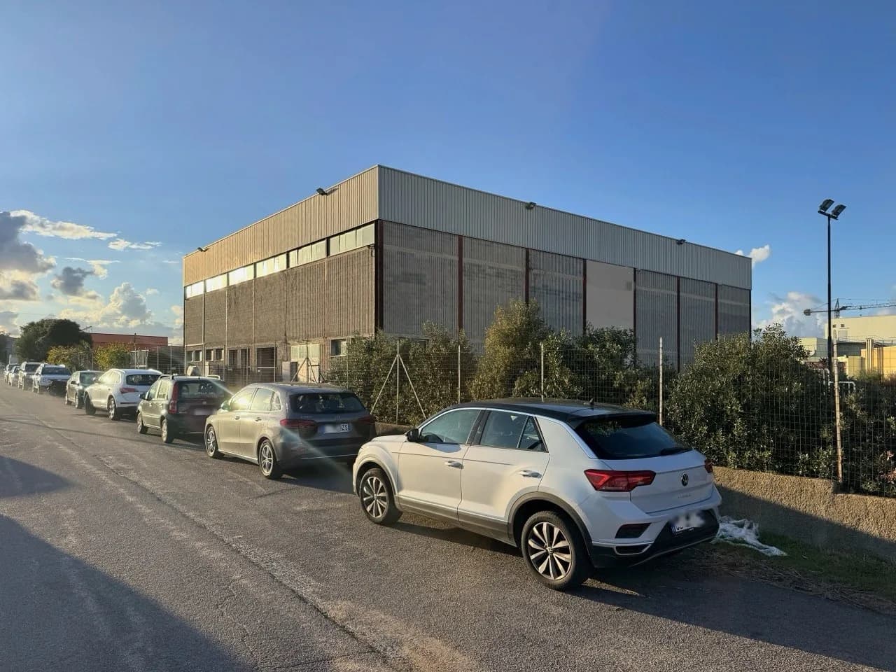 Grote, blokvormige sporthal in industriële stijl met metaalbeplating achter een draadhek en struiken, met auto's langs de weg onder een blauwe lucht in laat zonlicht