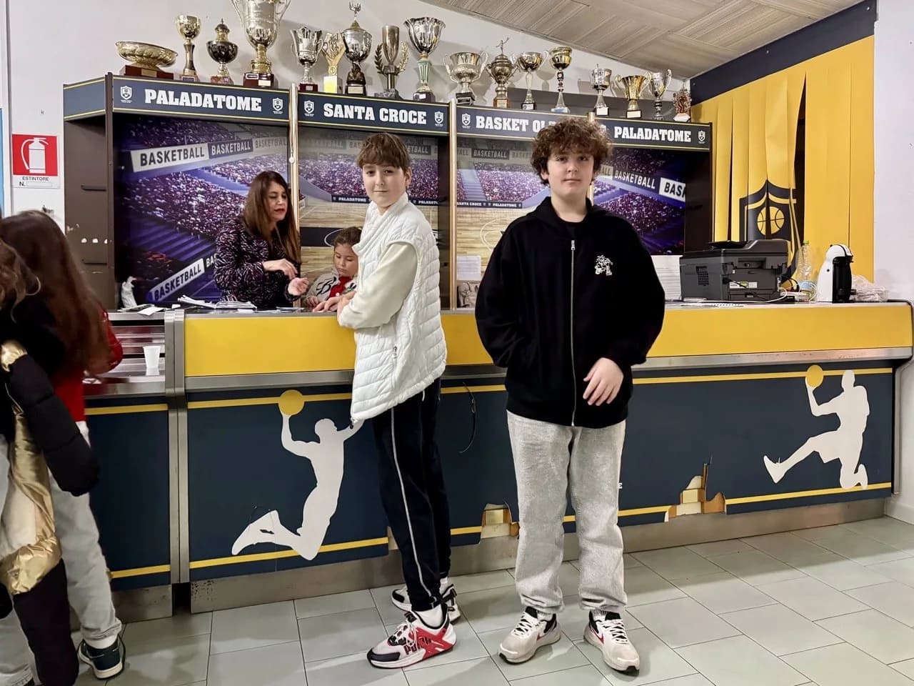 Twee jongens staan bij een geel-marineblauwe balie versierd met silhouetten van basketballers, met trofeeën op planken erboven en borden met Paladatome en Santa Croce.