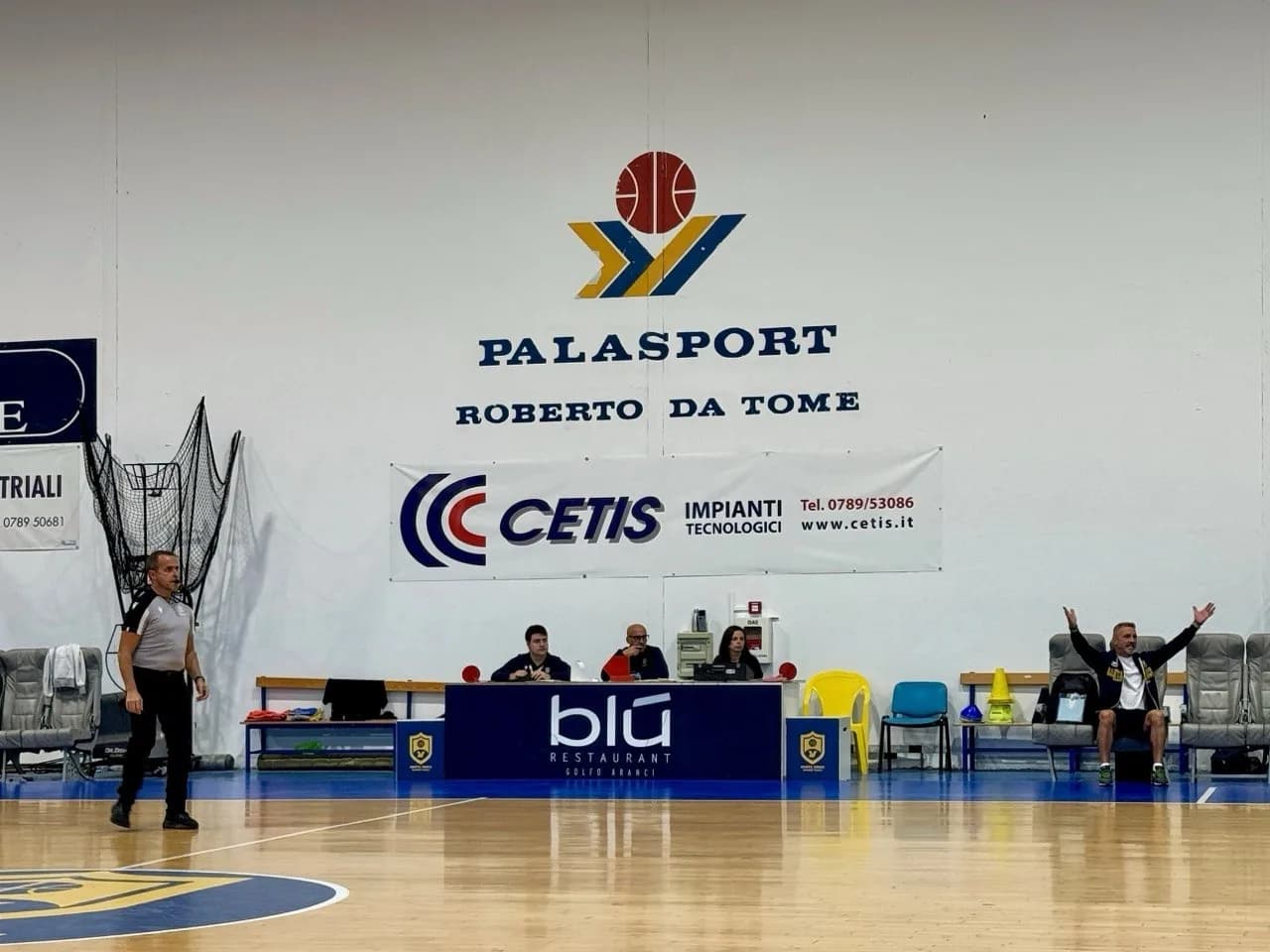 Overzicht van een overdekt basketbalveld met een houten vloer en blauwe randen, met spelers in geel en groen verzameld aan de overkant onder een stalen dak, een banner en scoreborden.