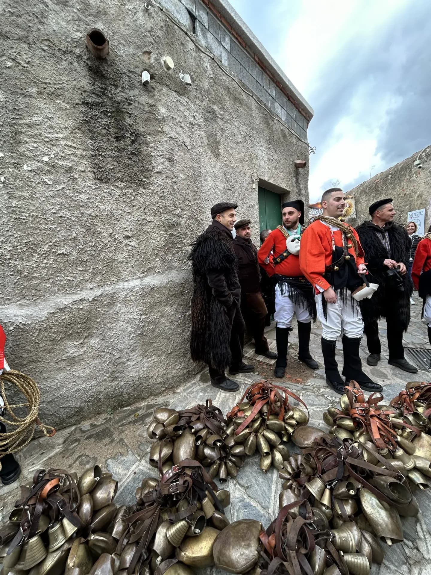 Men in traditional Sardinian costume, red jackets and sheepskins, standing beside piles of cowbells on a stone street