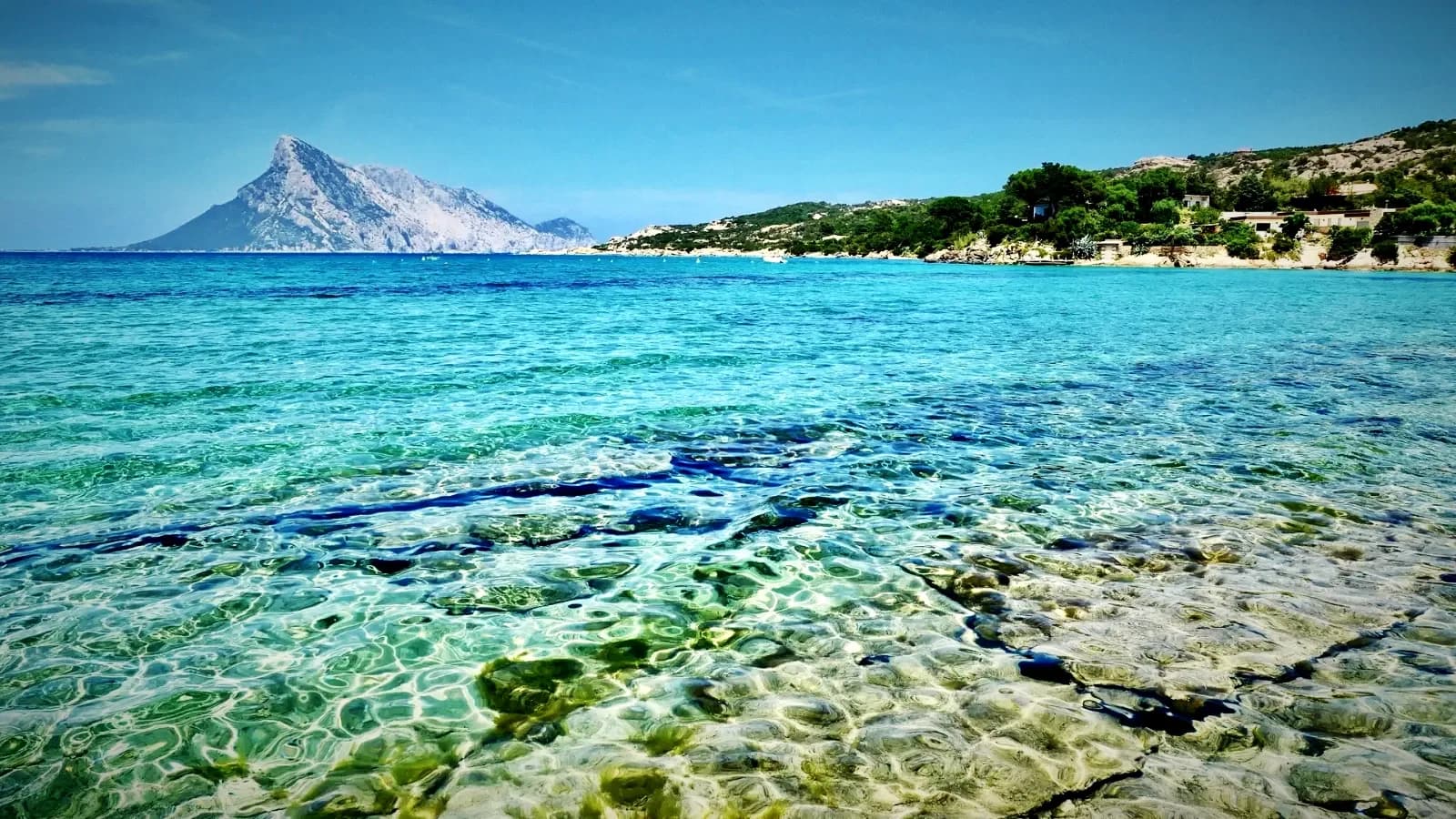 The island of Tavolara seen across clear shallow water from the shore at Porto San Paolo, with villas on the headland to the right, Gallura, Sardinia