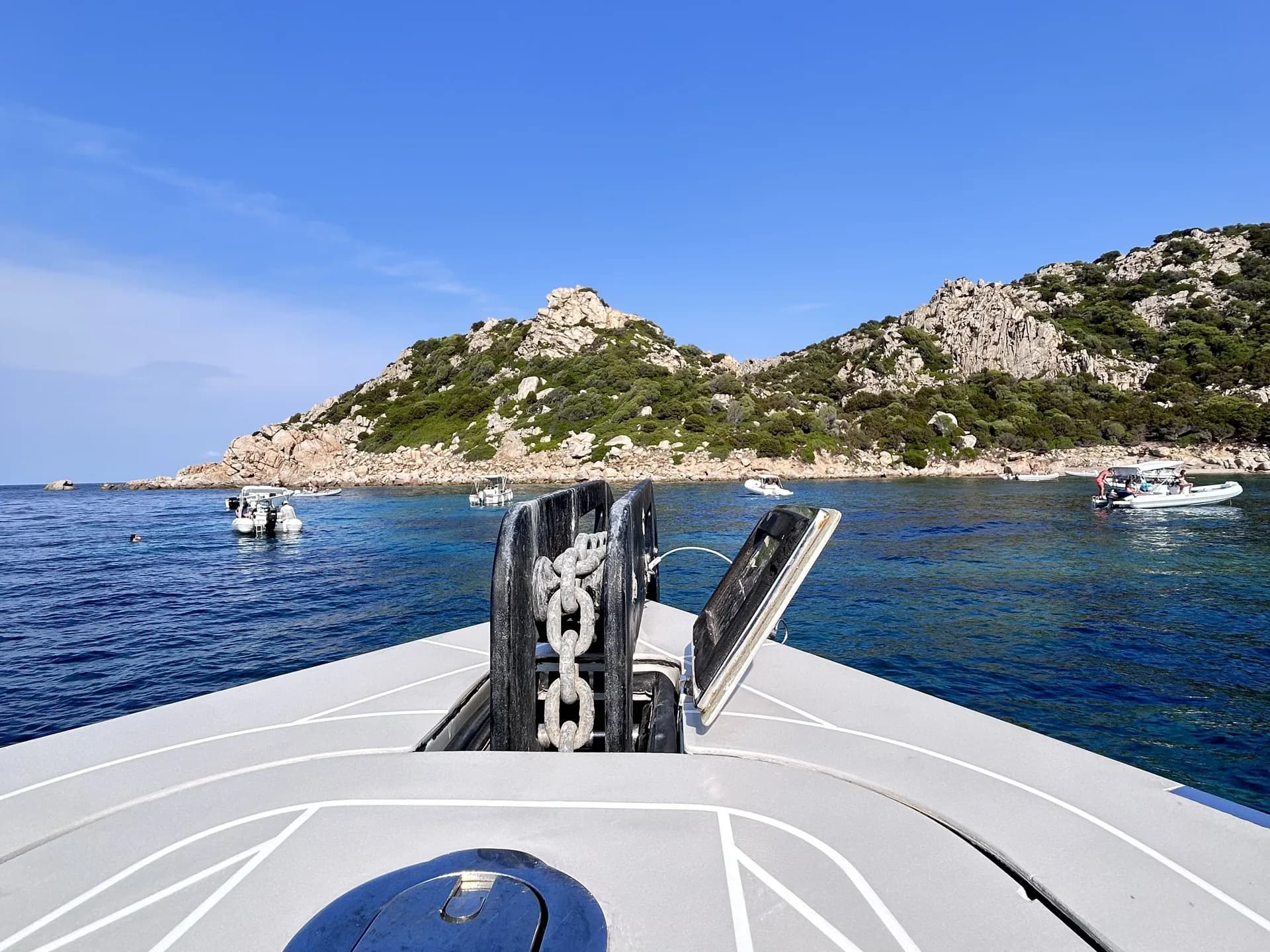 Vue depuis la proue d'un bateau, chaîne d'ancre visible, vers les pics calcaires escarpés de l'île de Tavolara et des bateaux amarrés dans une eau turquoise limpide.