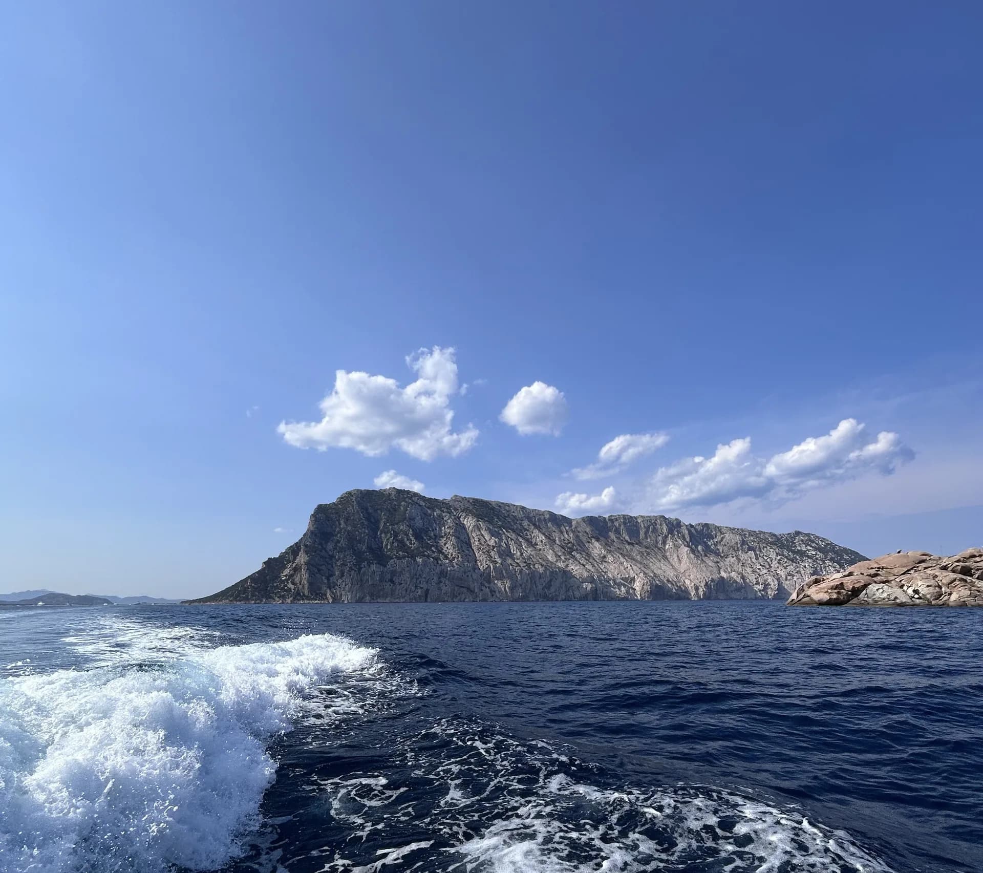 Le sillage blanc d'un bateau traverse la mer bleu foncé, tandis que les falaises calcaires escarpées de l'île de Tavolara s'élèvent sous un ciel lumineux parsemé de nuages.