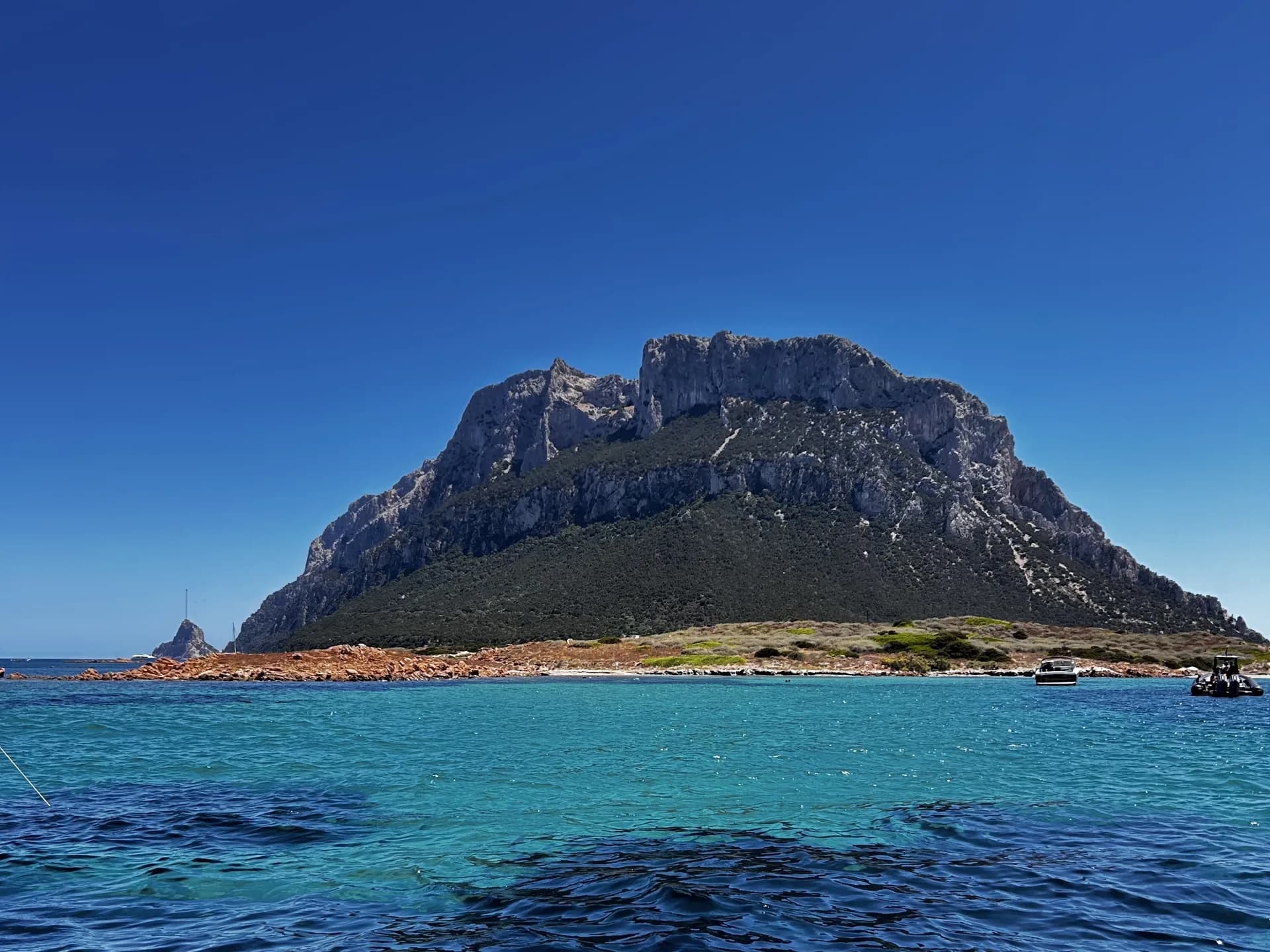 Le pic calcaire déchiqueté de l'île de Tavolara s'élève au-dessus d'eaux turquoise près d'Olbia, avec de petits bateaux ancrés le long de son rivage rocheux.