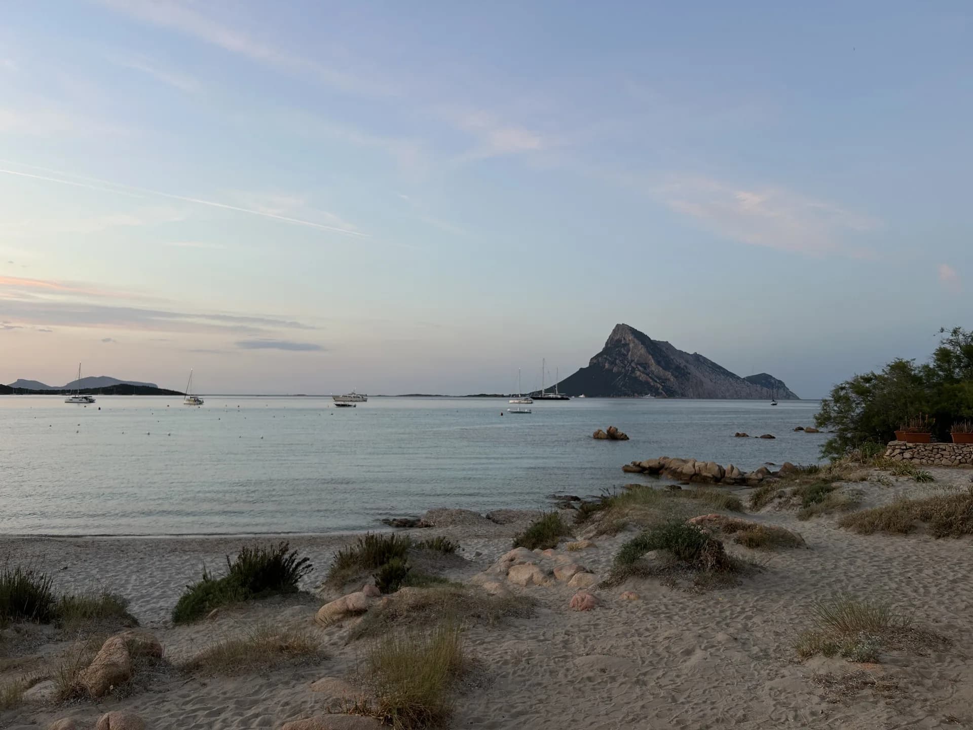 Vue depuis une plage de sable près d'Olbia, à travers une eau calme, vers le pic rocheux en forme de pyramide de l'île de Tavolara, avec des yachts ancrés au large au crépuscule.
