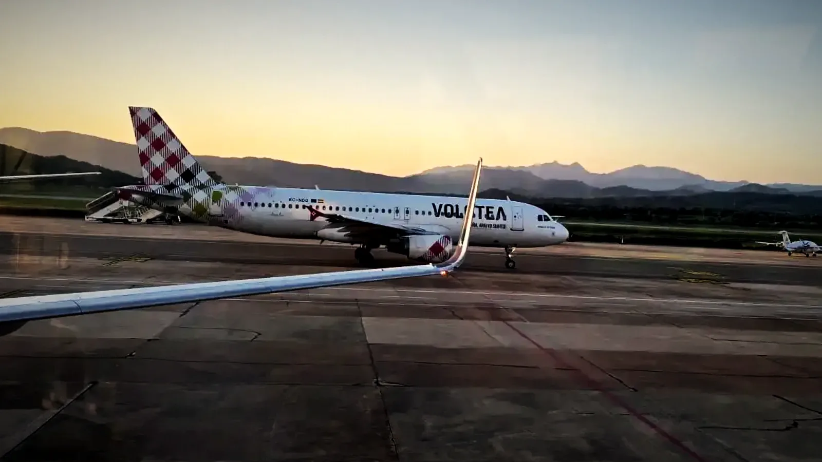 A Volotea Airbus A320 on the apron at Olbia Costa Smeralda airport at dusk, the Gallura hills behind, seen past an aircraft wing