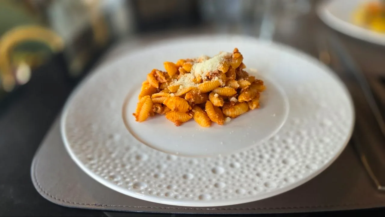 A serving of malloreddus pasta in tomato sauce, topped with grated cheese, on a white textured plate at a restaurant table.