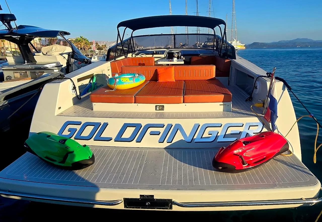 Stern of the yacht Goldfinger moored in a Sardinian marina, with green and red underwater sea scooters on the swim platform and orange cushioned seating on deck.