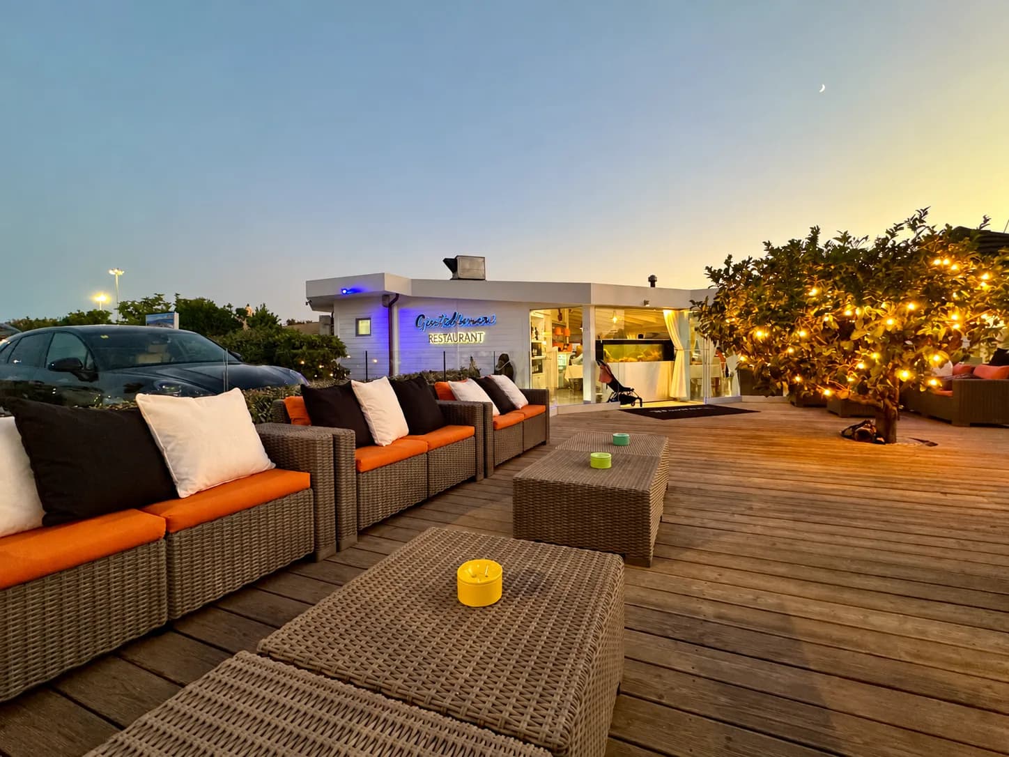 Wooden terrace at dusk with wicker sofas, orange cushions and low tables, a tree hung with fairy lights, and a glass-fronted restaurant with a lit sign.