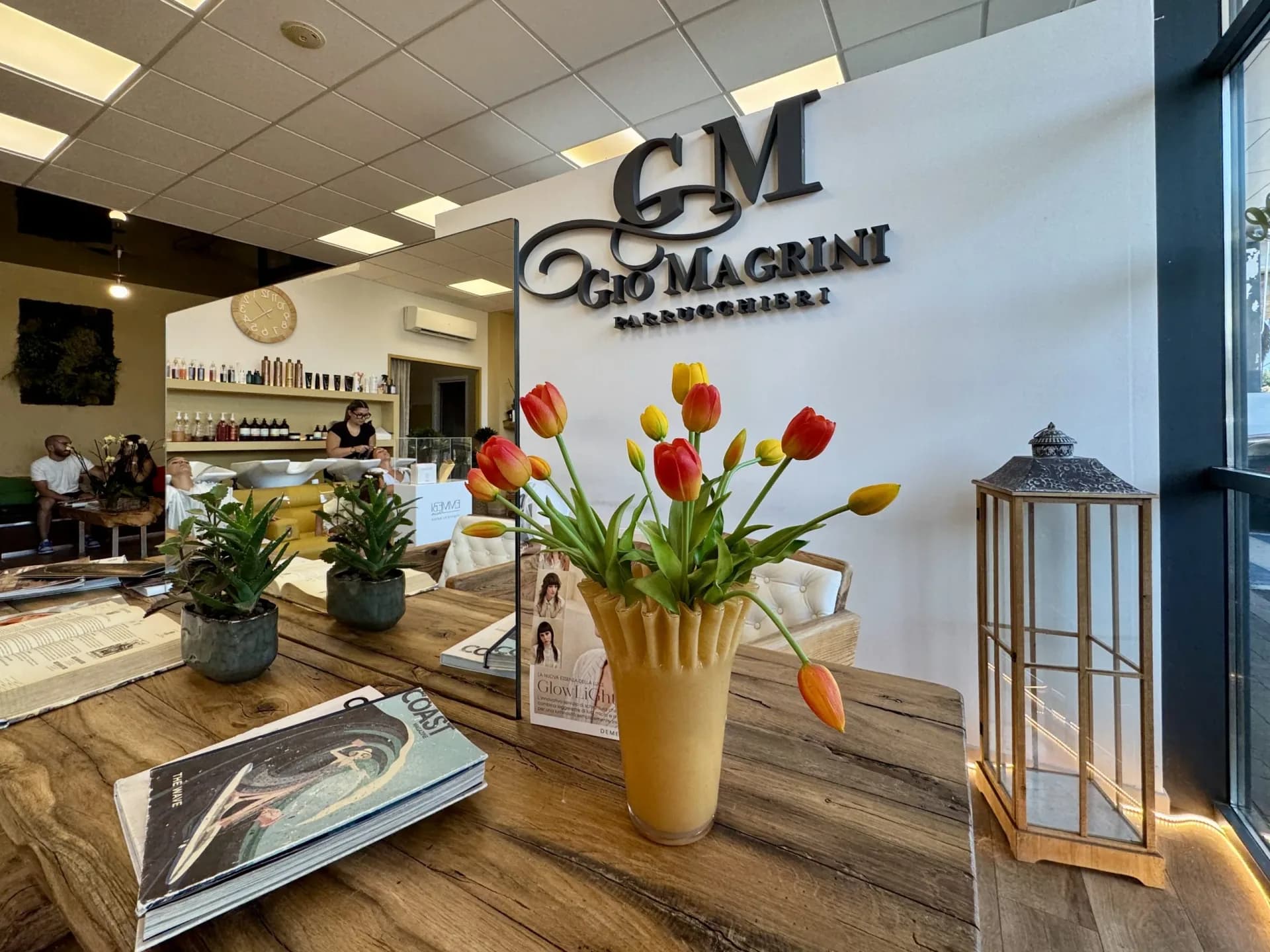 Gio Magrini Parrucchieri salon reception with wall logo, tulip vase, lantern and wooden table, backwash chairs visible in mirror behind.