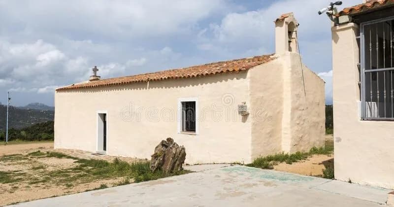 Whitewashed Chiesa Nostra Signora del Monte with terracotta roof tiles, bell tower and cross, set against Sardinian hills under a cloudy sky.