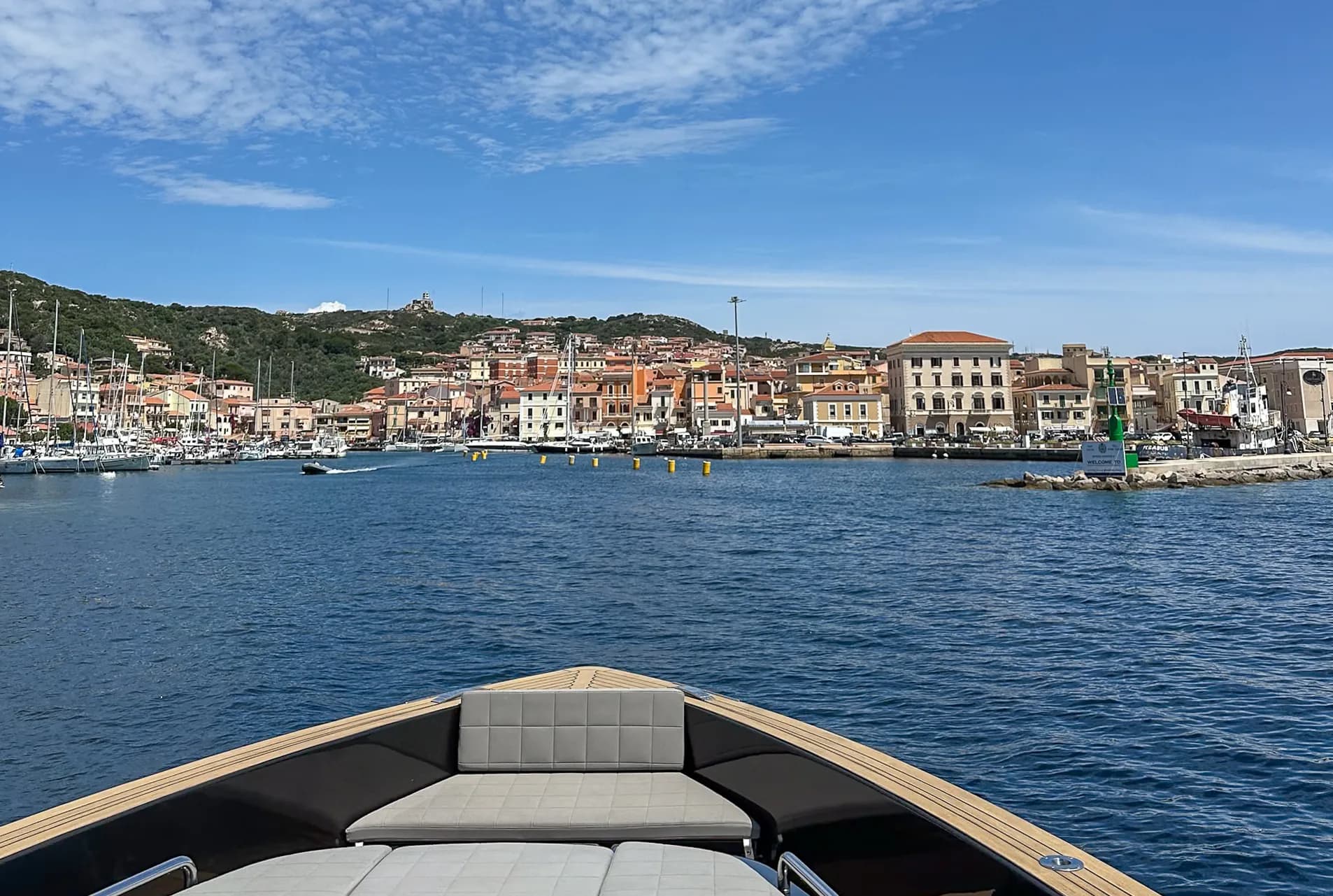 View from a boat's bow approaching La Maddalena's harbour, with moored yachts and colourful waterfront buildings beneath a hillside, Sardinia.