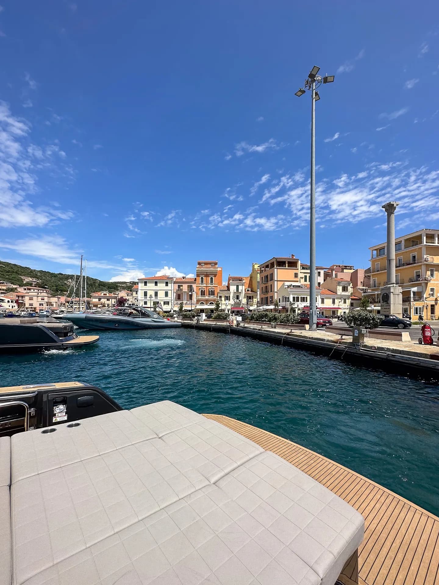 View from a yacht's deck across La Maddalena harbour, with pastel-coloured waterfront buildings, moored boats and clear blue water under a bright sky.