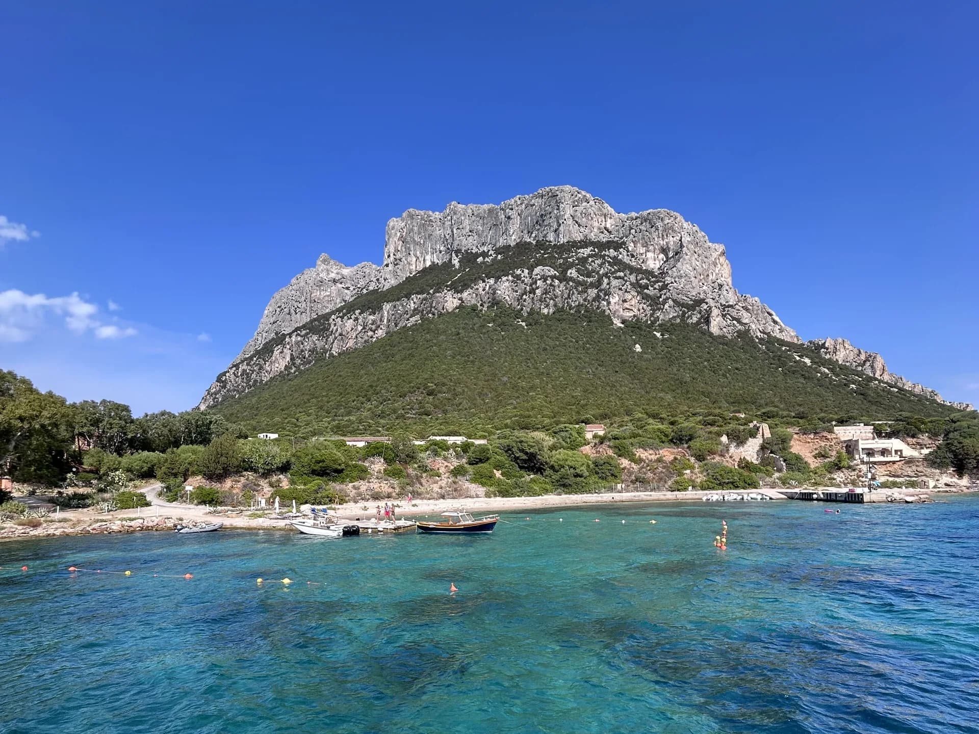 Tavolara Island's towering limestone peak rises above turquoise waters, with small boats moored near the shore and a cluster of buildings at its base.