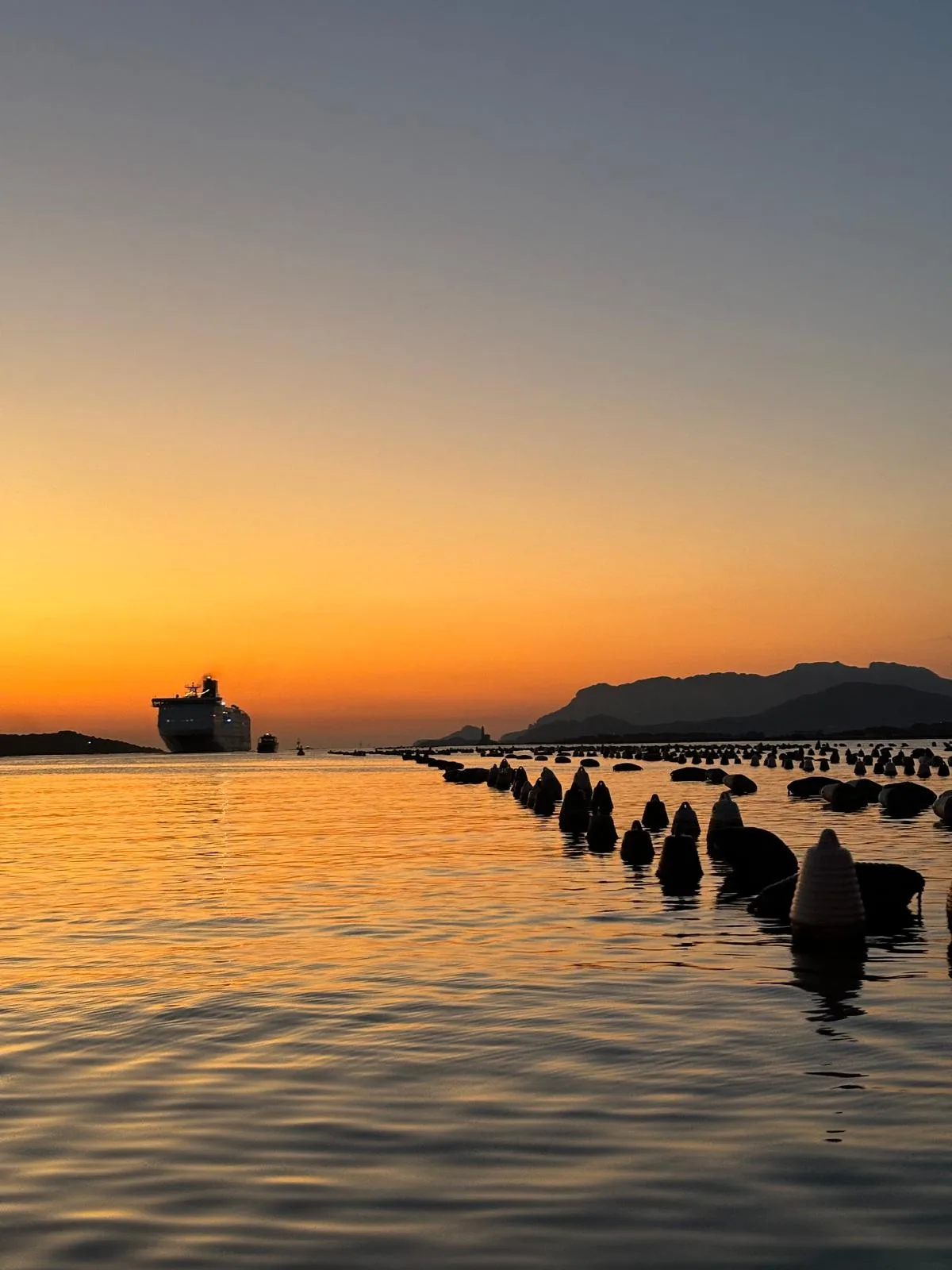 Sunset over the Golfo di Olbia, with a ferry on the horizon, rows of floats in the water and mountains in silhouette